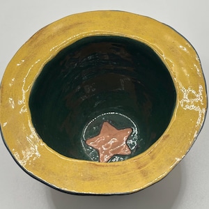 May include: A handmade ceramic bowl with a star design. The bowl has a dark green interior, a yellow rim, and a brown star at the bottom. The exterior is a dark blue.