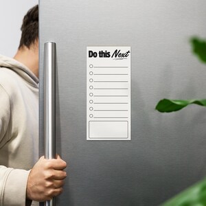 May include: A white rectangular magnet with the text "Do this Next" in black script. The magnet has a checklist format with circles and lines for notes, and is attached to a silver refrigerator door.