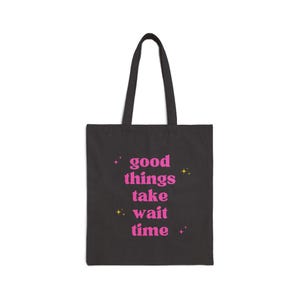 May include: Black tote bag featuring the pink text "good things take wait time". The bag has two black handles and is made of a durable fabric. The text is in a playful font, with star accents.