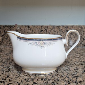 May include: A white porcelain gravy boat featuring a delicate floral pattern in soft pastel colors, accented by a navy blue and gold rim border. The vessel has a curved handle, a wide pouring spout, and a gold-trimmed base for stability.
