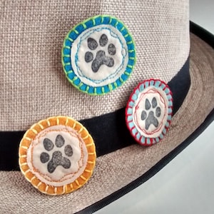 May include: Three fabric paw print patches with colorful stitching, attached to a brown hat with a black band.