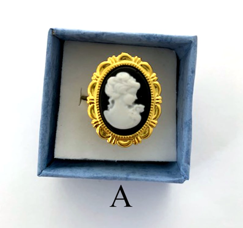May include: A gold ring with a black and white cameo of a woman's profile. The ring is in a blue velvet box with the letter "A" printed on the bottom.