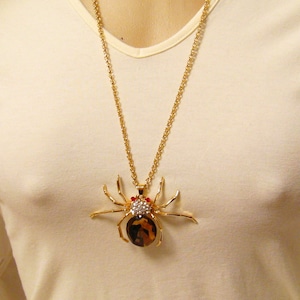May include: A gold chain necklace with a large, detailed spider pendant. The spider is made of gold metal and has red and clear rhinestones for eyes.
