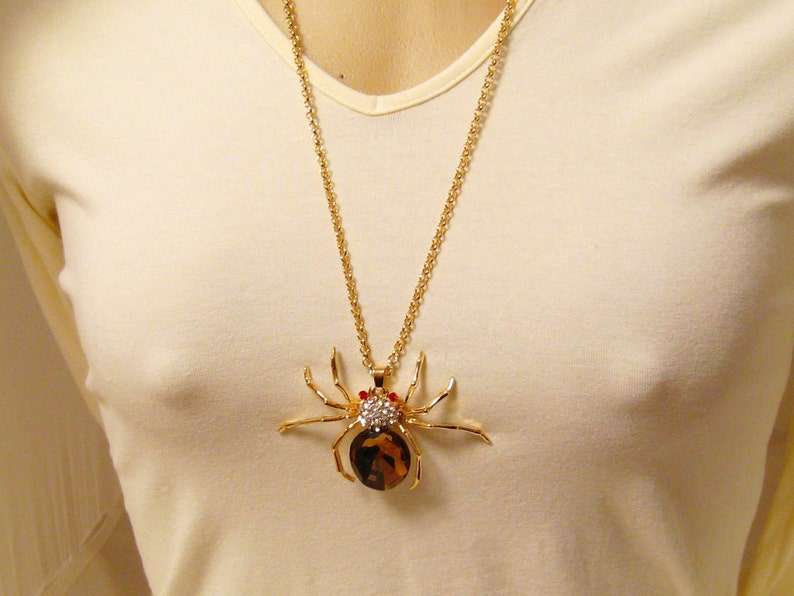 May include: A gold chain necklace with a large, detailed spider pendant. The spider is made of gold metal and has red and clear rhinestones for eyes.