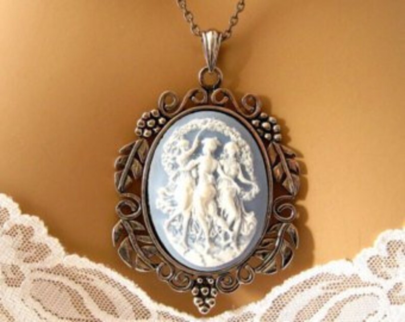 Op de afbeelding: Een zilverkleurige ketting met een hanger met een blauwe en witte cameo met drie vrouwen die in een cirkel dansen.
