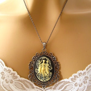 Op de afbeelding: Een zilveren ketting met een cameo-hanger met drie vrouwen in een zwart-wit ontwerp. De cameo is omgeven door een zilveren frame met bloemmotieven.