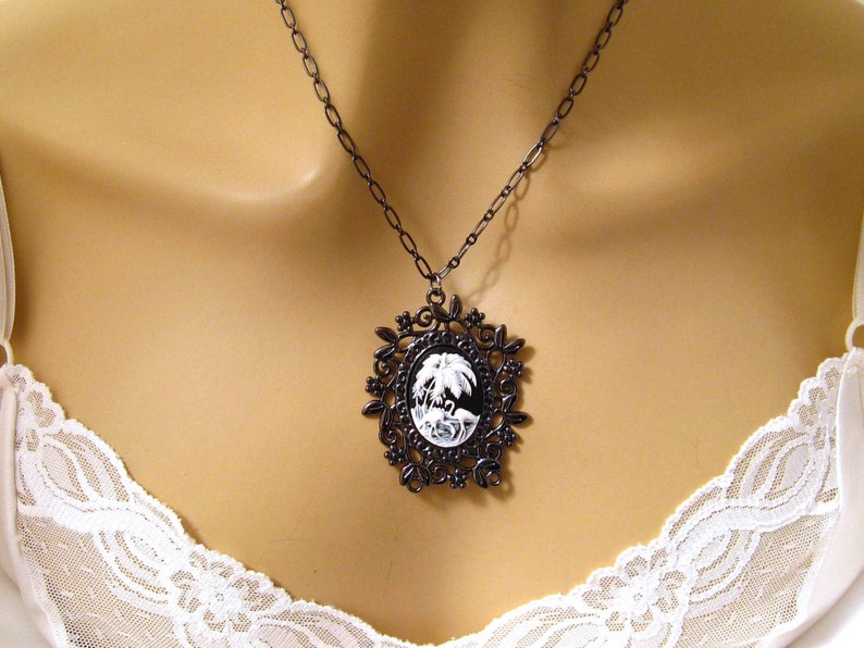 May include: A black cameo necklace with a white cameo featuring a palm tree and a bird. The cameo is set in a black filigree frame.