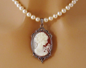 May include: A cameo pendant necklace featuring a profile of a woman with a flower, set in an ornate silver-tone frame. The pendant hangs from a strand of cream-colored faux pearls, creating a classic and elegant look.