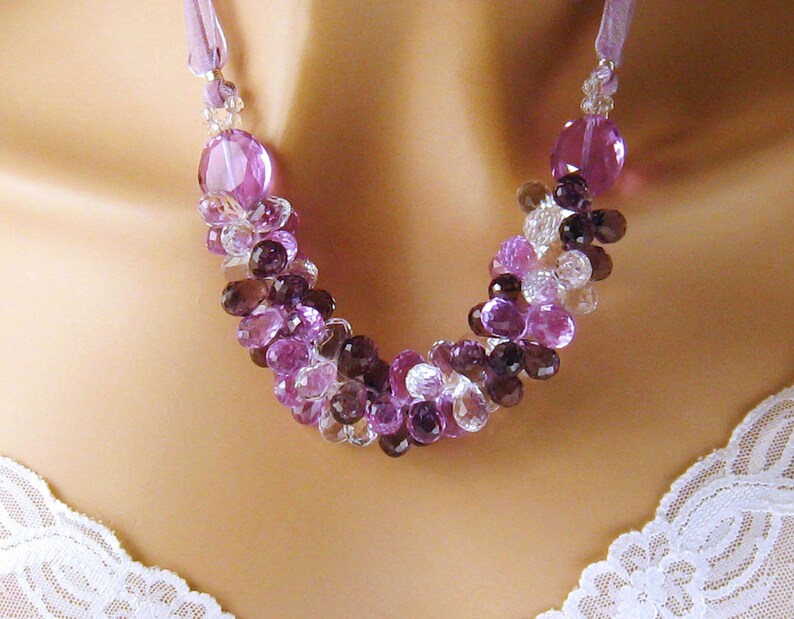 K&ouml;nnte beinhalten: Eine violette, rosafarbene und klare Kristallkette mit einem Satinbandverschluss. Die Kette besteht aus facettierten Edelsteinperlen in verschiedenen Violett- und Rosat&ouml;nen, wobei durchgehend klare Kristallperlen dazwischen liegen.