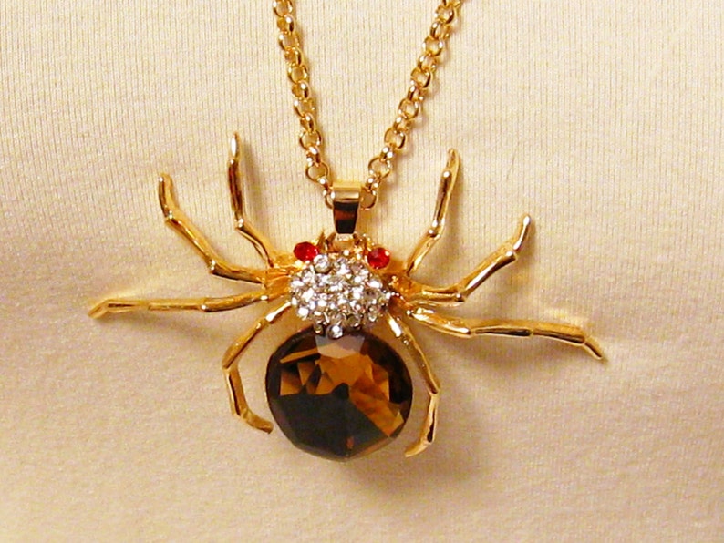 May include: A gold-tone spider pendant necklace with a brown crystal body and clear rhinestone accents. The spider has red eyes.
