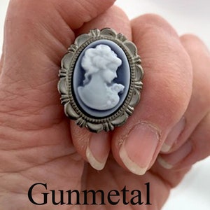 May include: A silver cameo ring with a white cameo of a woman's profile. The ring is set in a silver oval frame with a decorative border. The word "Gunmetal" is written below the ring.
