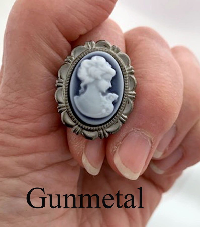 May include: A silver cameo ring with a white cameo of a woman's profile. The ring is set in a silver oval frame with a decorative border. The word "Gunmetal" is written below the ring.