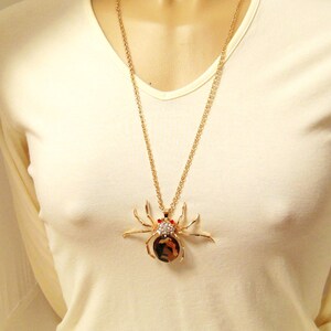 May include: A gold chain necklace with a large, detailed spider pendant. The spider is made of gold metal and has black and brown accents. The spider has red eyes and is covered in rhinestones.
