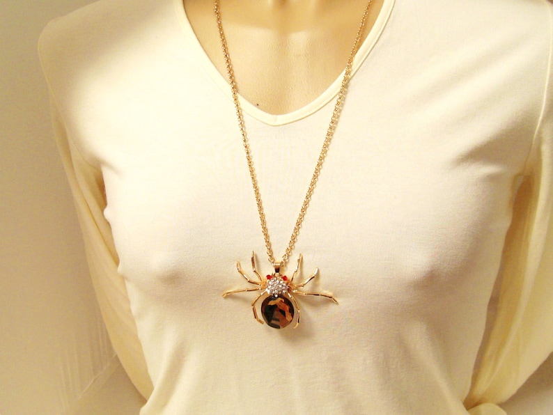 May include: A gold chain necklace with a large, detailed spider pendant. The spider is made of gold metal and has black and brown accents. The spider has red eyes and is covered in rhinestones.