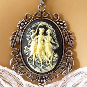 Op de afbeelding: Een zilveren ketting met een hanger met een cameo met drie vrouwen in zwart-wit. De cameo is gezet in een zilveren ovale lijst met een decoratief bloemmotief.