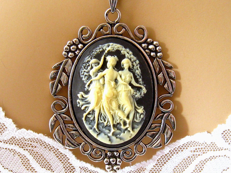 Op de afbeelding: Een zilveren ketting met een hanger met een cameo met drie vrouwen in zwart-wit. De cameo is gezet in een zilveren ovale lijst met een decoratief bloemmotief.