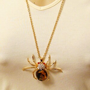 May include: A gold chain necklace with a large, detailed spider pendant. The spider is made of gold metal and has a brown gemstone body with clear and red rhinestones.