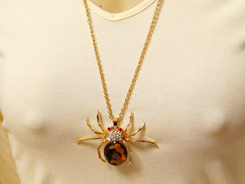 May include: A gold chain necklace with a large, detailed spider pendant. The spider is made of gold metal and has a brown gemstone body with clear and red rhinestones.