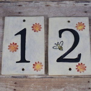 May include: Two white ceramic house numbers, one and two, with black numbers and orange flower accents. The number two also has a small bee painted on it.