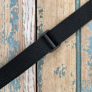 May include: Black adjustable strap with a plastic buckle. The strap is made of a woven fabric and is lying on a weathered wooden surface.
