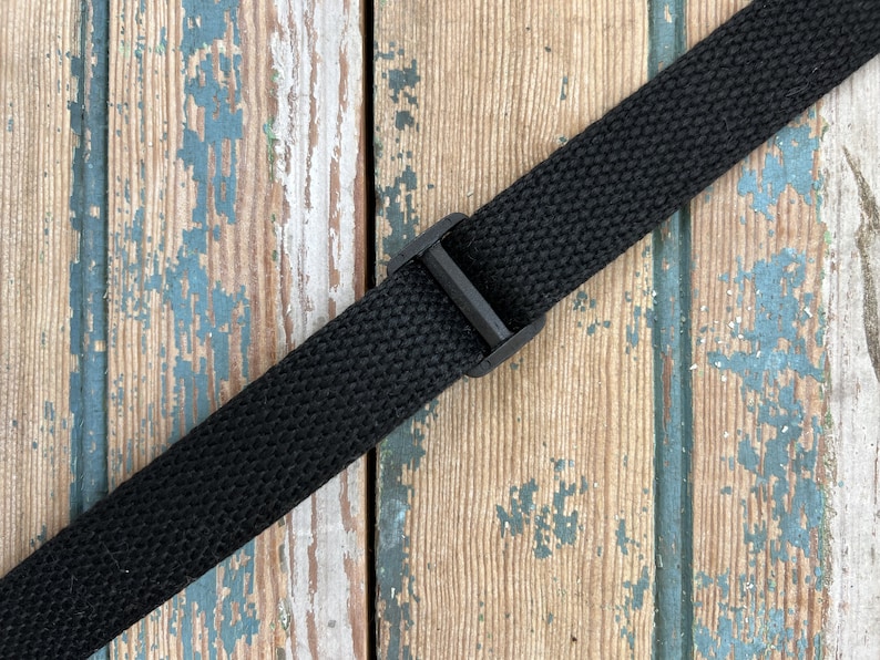 May include: Black adjustable strap with a plastic buckle. The strap is made of a woven fabric and is lying on a weathered wooden surface.