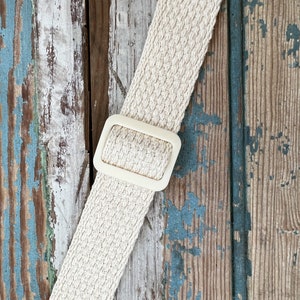 May include: A close-up of a beige woven strap with a white plastic buckle. The strap is against a weathered wooden background.