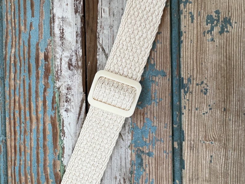 May include: A close-up of a beige woven strap with a white plastic buckle. The strap is against a weathered wooden background.