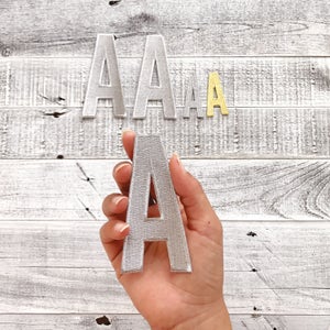 Könnte beinhalten: Drei silberne Filzbuchstaben-Patches in Form des Buchstabens A. Die Patches sind unterschiedlich groß. Der größte Patch wird in einer Hand gehalten.