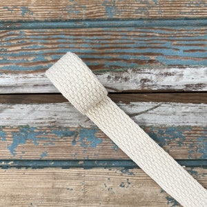 May include: A close-up of a natural off-white cotton webbing strap. The strap is lying on a weathered wooden surface with blue and white paint.