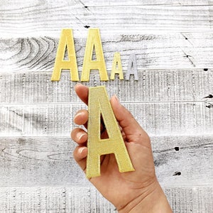 May include: Three gold felt letter patches in the shape of the letter A. The patches are different sizes. The largest patch is being held in a hand.