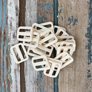 May include: A pile of off-white plastic slider buckles. The buckles are rectangular with two holes on each side.