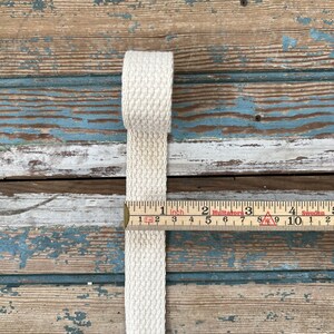 May include: A close-up of a natural-colored cotton webbing measuring 1 inch wide. The webbing is lying on a weathered wooden surface with a ruler measuring in inches.