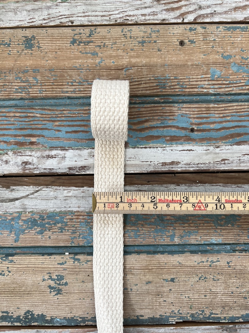 May include: A close-up of a natural-colored cotton webbing measuring 1 inch wide. The webbing is lying on a weathered wooden surface with a ruler measuring in inches.