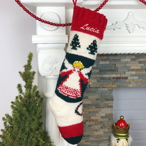 May include: A white, red, and green knitted Christmas stocking with an angel and Christmas trees design. The stocking is personalized with the name "Lucia".