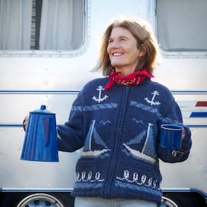 May include: A navy blue zip-up knit cardigan featuring a white and light blue sailboat and anchor pattern, worn with a red bandana. The garment is paired with light blue jeans, while holding a blue enamel coffee pot and a matching blue mug.