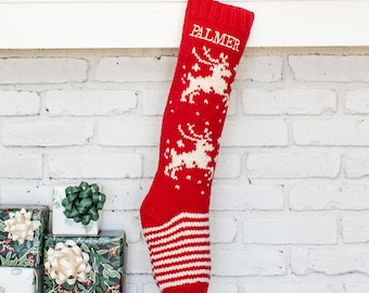 May include: A red knit Christmas stocking featuring the name PALMER in white capital letters at the top, two white reindeer silhouettes in the center, and a white and red striped pattern on the toe and heel area, hanging from a white mantel.