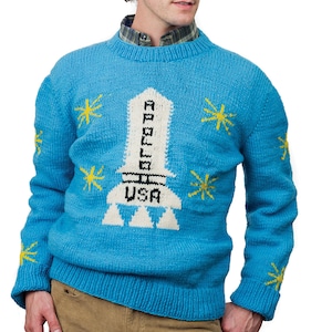 May include: A blue knitted sweater with a white rocket ship design and the words "Apollo" and "USA". The sweater has yellow stars on the sleeves and chest.