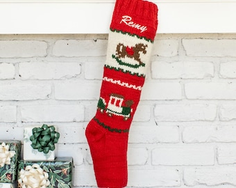 Puede incluir: Un calcetín navideño de punto en color rojo y crema con motivos de un caballo balancín y un tren. El nombre Remy está bordado en letra cursiva blanca en la parte superior. Tiene un puño acanalado y un pie rojo con la punta en crema.