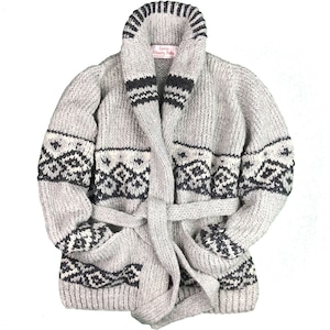 May include: A light gray, chunky knit cardigan with a shawl collar and a matching belt. The cardigan features black and white geometric patterns on the pockets and sleeves. The label reads "Camp Hilashby Knits".