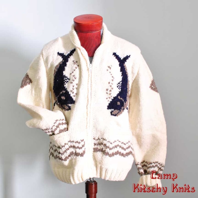 May include: Cream-colored zip-up sweater with a fish design. The sweater features two large fish in navy and brown on the front, with matching details on the sleeves and pockets. The sweater has a high collar and two front pockets.
