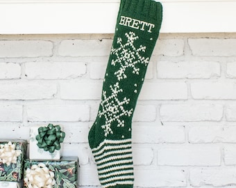 May include: A dark green knit Christmas stocking featuring a white snowflake pattern and the name BRETT embroidered in white at the top. The stocking has a ribbed cuff and a striped pattern at the toe, hanging vertically against a white brick wall.