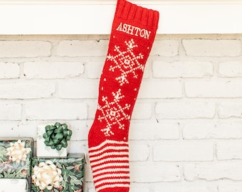 May include: A red knit Christmas stocking featuring a white snowflake pattern and the name ASHTON embroidered in white thread at the top. The stocking has a ribbed cuff and a white and red striped toe section, hanging from a small loop.