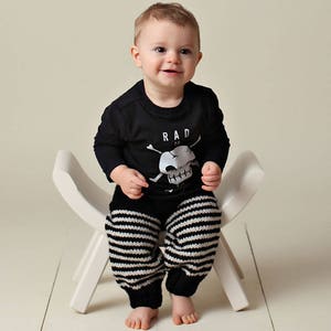 May include: A toddler wearing a black long-sleeved shirt with a white skull and crossbones graphic and the text "RAD" sitting on a white wooden stool. The toddler is wearing black and white striped pants.