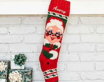 May include: A red knit Christmas stocking featuring a white, green, and peach-colored graphic of a person wearing glasses. The name Avery is embroidered in white cursive script at the top. The stocking has a ribbed cuff and a decorative patterned band.