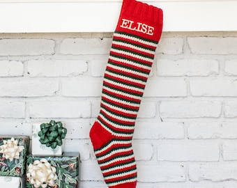 May include: A knitted Christmas stocking featuring horizontal stripes in red, white, and green. The top cuff is solid red with the name ELISE embroidered in white capital letters. The stocking includes a red hanging loop at the top edge.
