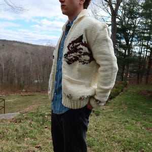 May include: Cream-colored knit cardigan with a brown and burgundy design on the front. The sweater has a shawl collar, pockets, and button closures. The person is wearing a denim shirt and black pants.