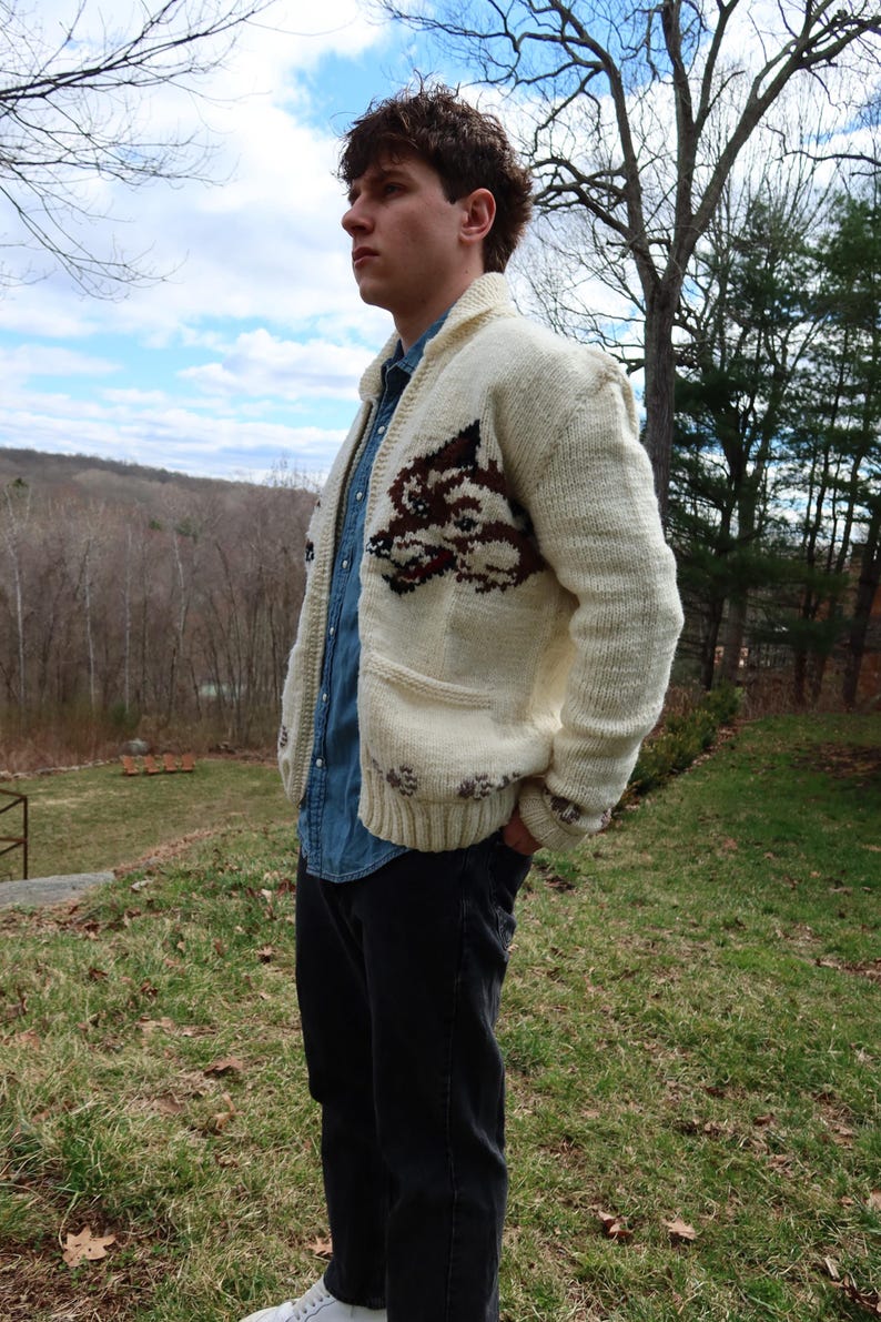 May include: Cream-colored knit cardigan with a brown and burgundy design on the front. The sweater has a shawl collar, pockets, and button closures. The person is wearing a denim shirt and black pants.