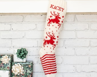 May include: A cream-colored knit Christmas stocking featuring red reindeer motifs and the name KACEY embroidered in red at the top. The foot section of the stocking displays a red and cream horizontal striped pattern throughout the lower portion.