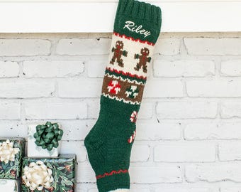 May include: A dark green knitted Christmas stocking featuring a white cuff with the name Riley embroidered in white script. The stocking displays a pattern of brown gingerbread men, red and white peppermint candies, and red and white candy canes.