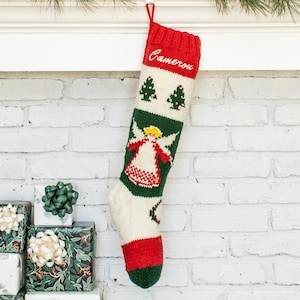 Puede incluir: Un calcetín navideño de punto con un puño rojo que tiene el nombre Cameron bordado en blanco. El calcetín presenta un patrón de dos árboles de Navidad verdes, una figura central de un ángel con alas blancas y una puntera y talón rojos.
